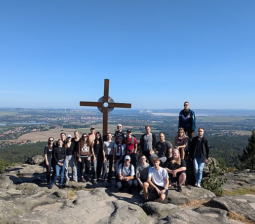 Ein Gruppenbild von einer studentischen Wandertruppe im Zittauer Gebirge vor einem Gipfelkreuz unter blauem Himmel.