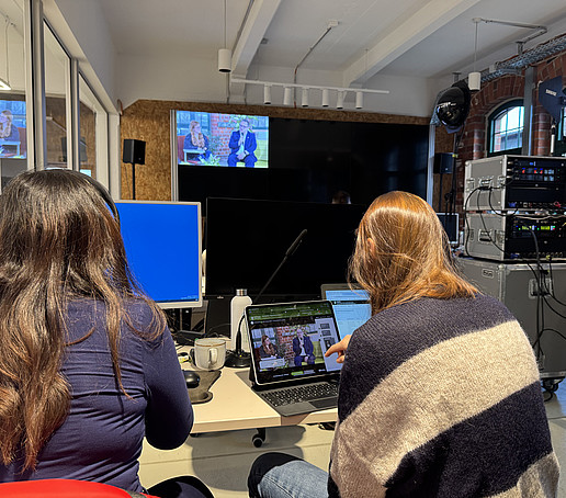 In einem vom Studio mit Glaswand abgetrennten Raum sitzen zwei Mitarbeiterinnen vor Bildschirmen und schauen darauf. Im Hintergrund ist ein großer Screen zu sehen.