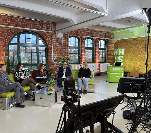 In einem Studio mit Backsteinmauern und großen Fenstern im Hintergrund sitzen Teilnehmende auf grünen Sofas verteilt. Im Vordergrund stehen schwarze Bildschirme auf dem Boden. Am Rand steht ein Beleuchtungsschirm.