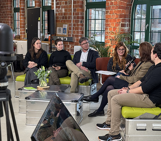 In einem Studio mit Backsteinmauern und großen Fenstern im Hintergrund sitzen Teilnehmende auf grünen Sofas verteilt und unterhalten sich.