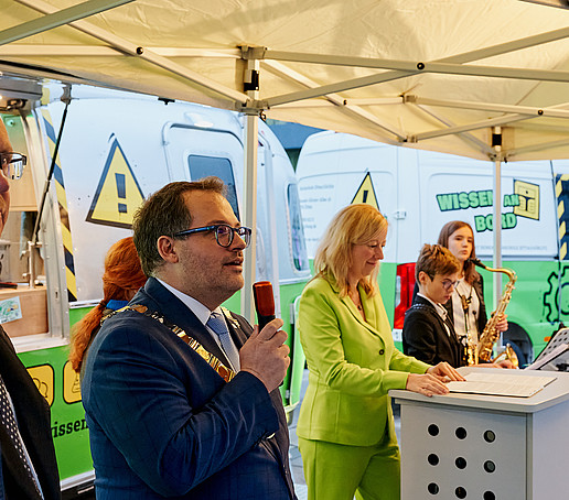 Der Rektor spricht ins Mikro, neben ihn der Hochschulratsvorsitzende, die Prorektorin und junge Blechbläser. Sie stehen vor dem ZukLOS-Anhänger.