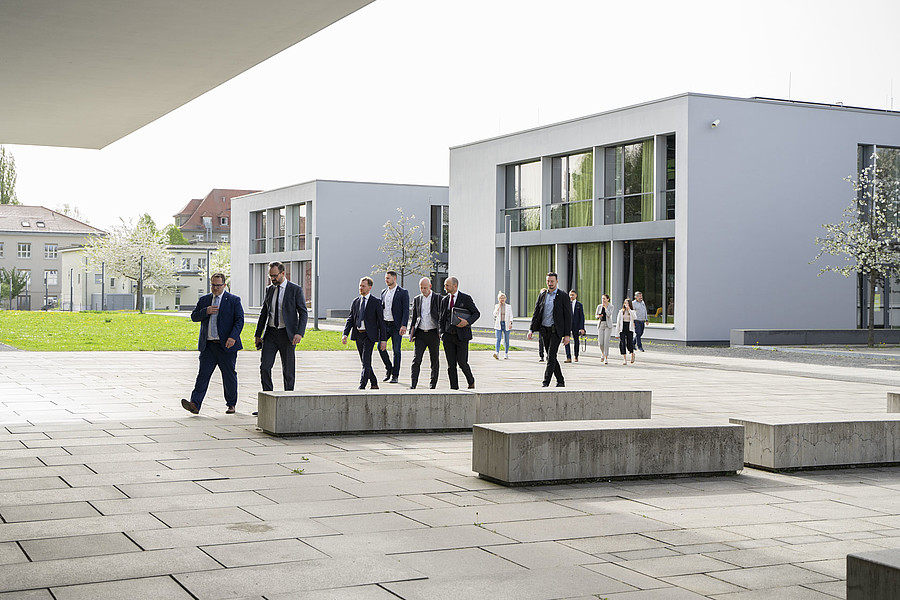 Gruppe von formell gekleideten Personen geht durch einen modernen Campus.