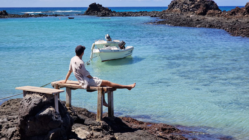 A young man sits on a bench on the beach. A white boat floats in the bay on the azure blue water in front of him.