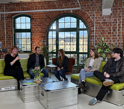 In einem Studio mit Backsteinmauern und großen Fenstern im Hintergrund sitzen Teilnehmende auf grünen Sofas verteilt und unterhalten sich.