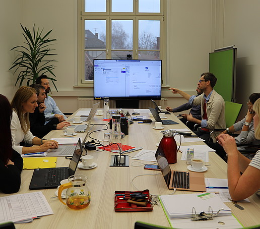 In einem Sitzungssaal sind Mitarbeitende versammelt, die mithilfe eines Bildschirms das Thema DMS besprechen.