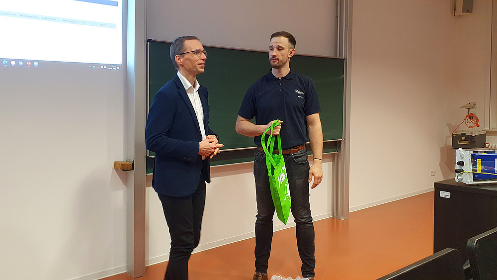 Prof. Kornhuber und Herr Schumann stehen vor der Tafel im Vorlesungssaal. Herr Schumann hält einen grünen Beutel in der Hand, während Prof. Kornhuber spricht.