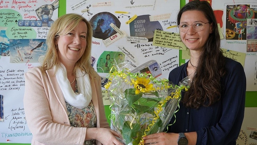 Die Prorektorin und Tanja Schwarz halten vor einer Pinnwand einen Blumenstrauß zwischen sich in den Händen.