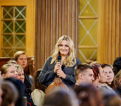 Eine Studentin hält ein Mikro in der Hand und hat sich aus einer vollbesetzten Sitzreihe erhoben.