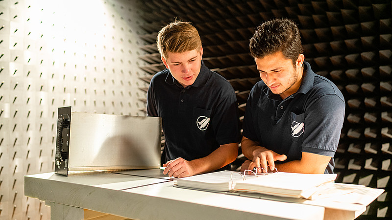 Zwei KIA-Studenten blättern in einem offenen Ordner und stehen im Energietechniklabor.