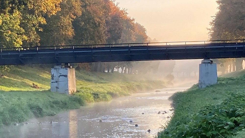 Eine Brücke führt über einen Fluss im Hintergrund herbstliche Bäume.