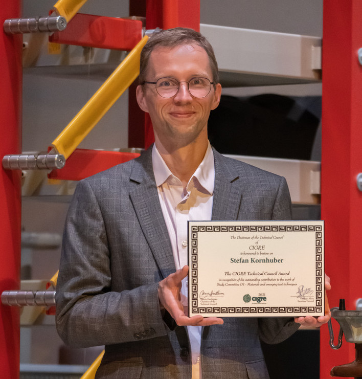 Prof. Kornhuber hält seine CIGRE-Award-Urkunde in die Kamera. Er steht in der Hochspannungshalle der HSZG.