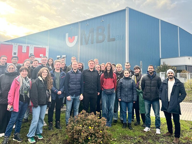 A group of students are standing on the premises of a factory.