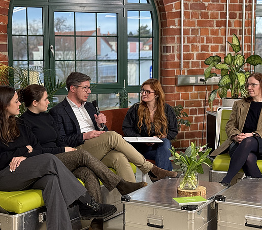 In einem Studio mit Backsteinmauern und großen Fenstern im Hintergrund sitzen Teilnehmende auf grünen Sofas verteilt und unterhalten sich.