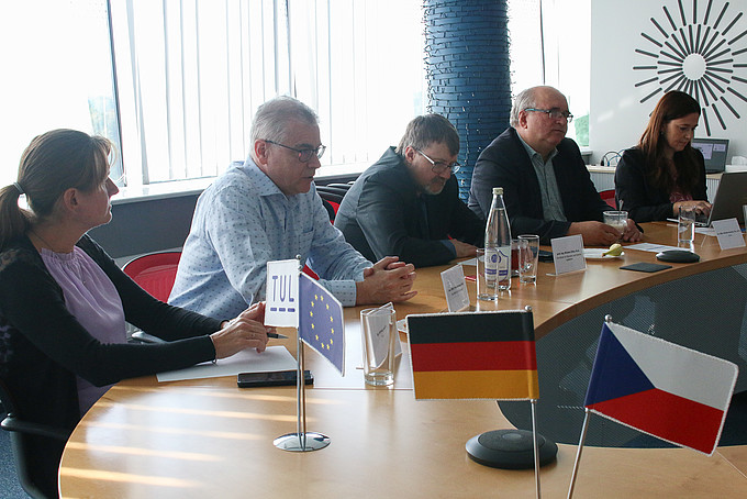 Drei Männer und zwei Frauen sitzen an einem Tisch. An der Tischseite im Vordergrund sind die deutsche und die tschechische Flagge als Tischschmuck aufgestellt.