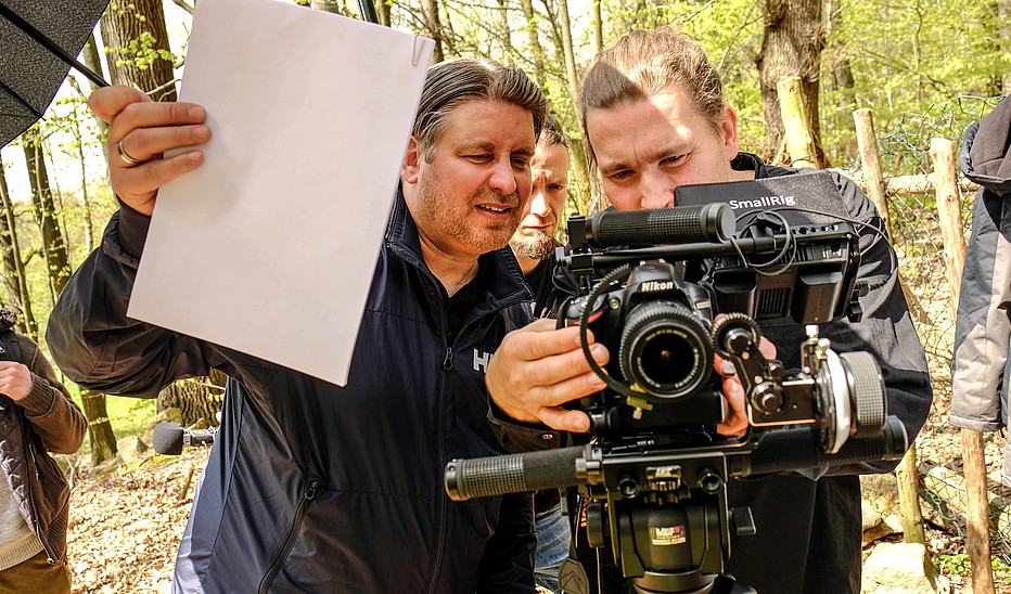 Ein Kameramann steht im Wald hinter einer Kamera und stellt diese ein. Ein Assistent hält ein weißes Blatt zum Weißabgleich in der Hand.