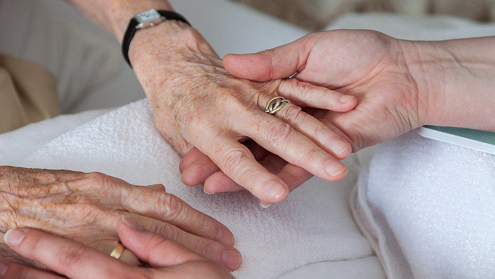 Two hands of a clearly older person hold the hands of a younger person.