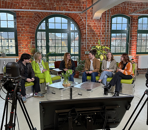 In einem Studio mit Backsteinmauern und großen Fenstern im Hintergrund sitzen Teilnehmende auf grünen Sofas verteilt und unterhalten sich.