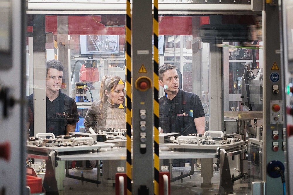 Three people observe a production plant behind a pane of glass.
