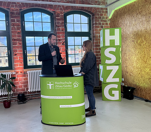 Ein Mitwirkender und die Moderatorin stehen im Studio hinter einem HSZG-Theke. Links im Vordergrund steht eine schwarze Kamera. Rechts von der Theke sind vier HSZG-Würfel aufgetürmt.