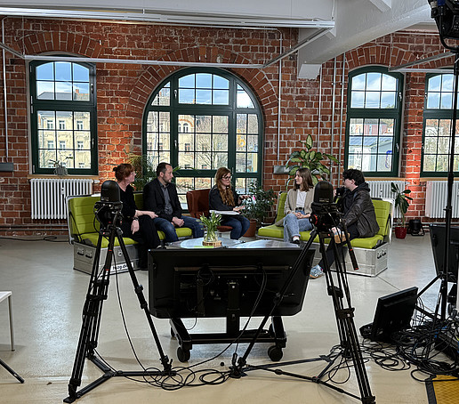 In einem Studio mit Backsteinmauern und großen Fenstern im Hintergrund sitzen Teilnehmende auf grünen Sofas verteilt und unterhalten sich.