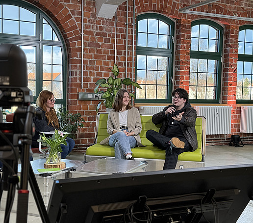 Zwei Teilnehmende sitzen auf einem grünen Sofa. Der rechte spricht in ein Handmikrofon.